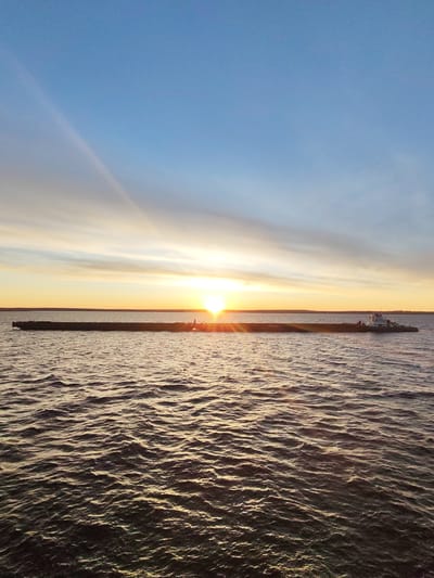 The image depicts a body of water, possibly a river or a large lake, at sunset or sunrise. A long, low barge is visible in the center of the frame, stretching across the horizon line. A tugboat is attached to the barge on the right side. The sun is positioned directly behind the barge, causing a silhouette effect and creating sunbeams that extend into the sky. The water's surface is rippled, reflecting the golden light of the sun. The sky transitions from a light blue at the top to a gradient of orange and yellow near the horizon. No people are directly visible, although they may be on the boat. Maksakovo, Russia, is the specified location. The environment is aquatic and characterized by a natural sunset scene.