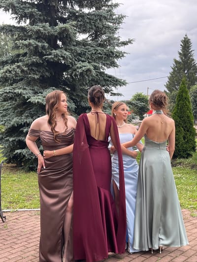 Four young women are standing in a circle, facing away from the camera, in what appears to be a garden setting in Starobaltachevo, Russia. They are dressed in formal, floor-length gowns. The woman on the far left wears a brown off-the-shoulder dress with a slit up the leg. Next to her, a woman in a deep red dress with a cape-like overlay. The third woman from the left is in a light blue off-the-shoulder dress. The woman on the far right is in a pale green halter dress. They are positioned in front of a large evergreen tree, with a smaller evergreen tree and other foliage visible in the background. The ground is paved with red bricks. The sky is overcast.