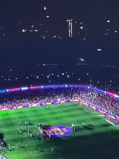 The image depicts a night scene, possibly of a sporting event or concert. A large crowd of people encircles a brightly lit green field. The edges of the field are illuminated with colorful lights, likely decorative or for the event's staging. In the background, there are clusters of lights, suggesting a city or urban area. Without further detail in the image or external knowledge, it is not possible to confirm the location of Sant Martí Sarroca or the specifics of the event.