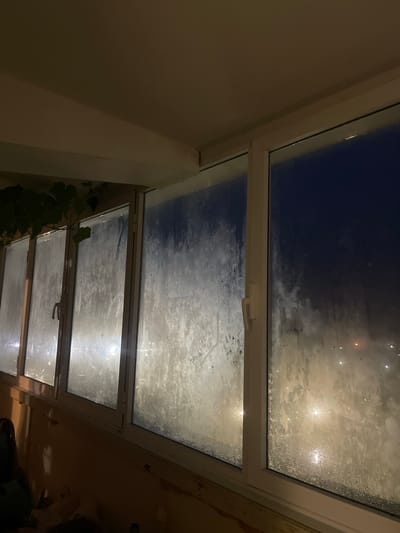 A nighttime, low-angle shot captures a condensation-covered window overlooking a city at night. The window is divided into several panes, with the left-most panes showing a more diffuse view of lights, while the right-most pane offers a clearer, darker view of the city's expanse. The glass is heavily fogged, obscuring details and creating a hazy effect. Streaks and droplets of condensation are visible across the surface. The interior side of the window frame is visible, along with a portion of the ceiling and wall. A plant with dark green leaves hangs from the upper left corner, partially obscuring the window. The scene appears to be from an apartment in Laishevo, Russia, given the context.
