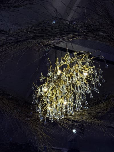A close-up, low-angle shot captures an ornate chandelier hanging from a dark, textured ceiling. The chandelier is made of golden, branch-like structures from which numerous clear, teardrop-shaped glass ornaments are suspended. Several small lights are visible within the chandelier, illuminating the glass droplets and casting a warm glow. The ceiling is adorned with a dense arrangement of thin, dark branches, creating a naturalistic and somewhat whimsical atmosphere. The overall lighting is dim, with the chandelier being the primary light source, contrasting with the dark, twig-covered environment. The scene appears to be an interior space, possibly a restaurant or event venue, in Bakhtiyary, Russia.