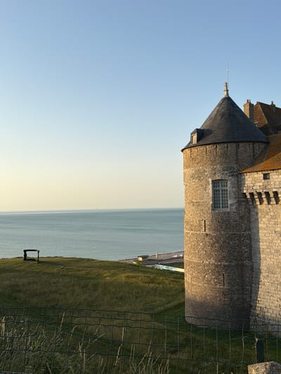 This is a photograph taken in Dieppe, France, of a building with a conical roof, possibly part of a historic structure like a lighthouse or a castle. The building is constructed of stone blocks and has a window with a grid pattern. Adjacent to the building is a body of water, likely the sea, stretching out to the horizon under a clear blue sky that transitions to a pale yellow near the horizon, suggesting it is either early morning or late afternoon.
The foreground shows a grassy slope leading down to the water, with a wire fence running across it. There's a simple, dark-colored object, possibly a bench or part of a railing, visible on the grass. The overall scene evokes a sense of tranquility and historical atmosphere. The light suggests a calm weather day. There is no visible text in the image.
