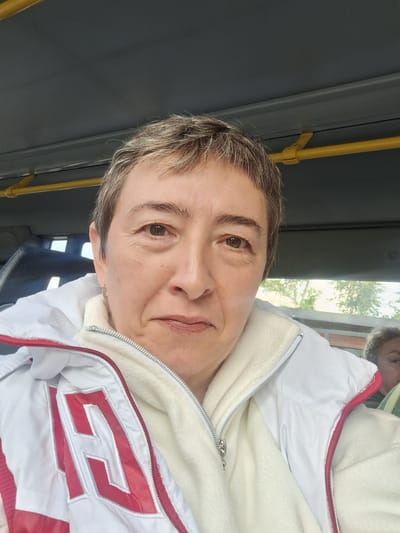 A woman, appearing to be middle-aged, is taking a selfie on what seems to be a bus in Gorka-Pokrovskaya, Russia. She has short, graying hair and is looking directly at the camera. She is wearing a white jacket with red trim and a white fleece underlayer. A yellow pole is visible above her head. To the right and slightly behind her, another person's head and shoulders are visible. The background suggests an outdoor environment with trees and buildings visible through a window.