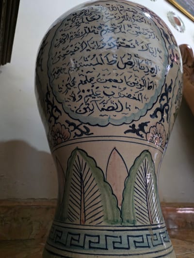 The image shows a close-up of a decorative ceramic vase with Arabic calligraphy. The vase is primarily off-white with blue and green painted details. The calligraphy, in dark blue, covers a significant portion of the upper section of the vase, arranged within a cloud-like border. Below this, there are floral motifs and stylized green leaf designs, followed by a geometric pattern along the base. The vase appears to be an interior decorative object. No specific location in Bireuenmeunasahblang, Indonesia can be identified from the provided image.