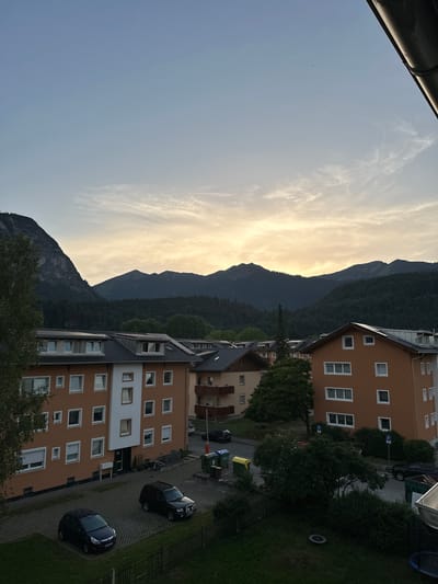 This is a vertically oriented, eye-level shot of a residential area with mountains in the background. The scene depicts a residential building with an orange facade, white-framed windows, and dark gray roofing. Several other similar buildings are visible. A paved parking lot runs alongside one of the buildings, with two cars parked. Green grass and trees surround the area. The sky is a pale blue with wispy clouds catching the golden light of a setting sun. The overall mood is peaceful and serene. The image appears to have been taken in the late afternoon or early evening, with the sun casting long shadows and bathing the scene in warm light. The presence of the mountains suggests a location in a mountainous region, consistent with Garmisch-Partenkirchen, Germany. There is a parking sign visible on one of the buildings.