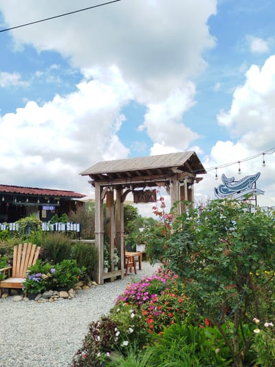 This is an outdoor scene in the city of Xuan Truong - Da Lat, Vietnam, featuring a garden cafe. The foreground is dominated by a gravel path and abundant flowering plants in vibrant pink, red, and purple hues, along with lush green foliage. To the left, a light-colored wooden Adirondack chair sits near a potted plant with purple flowers.
A rustic wooden archway stands in the mid-ground, adorned with string lights. Behind the archway, a building with a dark red roof is visible, featuring a sign that reads "ORDER HERE!". Further to the right, a large blue graphic of a whale-like creature is displayed against the sky, also illuminated by string lights. The sky above is a clear blue with scattered white cumulus clouds, and a thin black wire runs across the top of the frame. The overall atmosphere suggests a tranquil and picturesque outdoor dining or relaxation space.