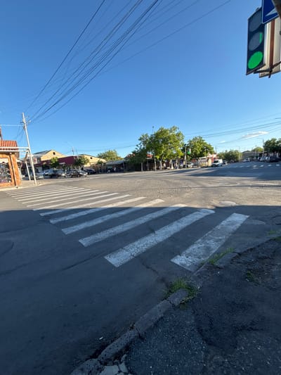 The image shows a street scene in Ashtarak, Armenia. The perspective is from a low angle, looking up at a clear blue sky, with power lines crisscrossing the frame. On the left, a traffic light shows a green signal. The street itself is paved and has a pedestrian crossing marked with white stripes.

Along the right side of the street, the asphalt is cracked and uneven, with patches of grass growing through. On the left side of the street, there are buildings, trees, and parked cars. The architecture of the buildings suggests a residential or commercial area.

The time of day appears to be daytime, likely late morning or early afternoon, given the bright sunlight and clear sky. The weather seems to be warm and sunny.

The scene depicts a typical street in a town or city, with infrastructure like traffic lights and power lines, as well as everyday elements like buildings and vehicles. There are no people clearly visible engaging in any specific activity, but the presence of parked cars and buildings suggests an inhabited area.