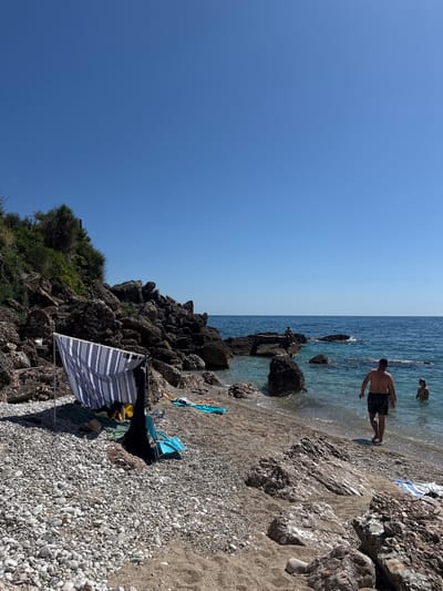 This image depicts a sunny day on a rocky and pebbly beach in Drobnići, Montenegro. The scene is dominated by a clear blue sky, featuring a faint white contrail in the upper mid-right, and a calm, turquoise-blue sea.
On the left side of the beach, a blue and white striped windbreak is erected, providing shade. Beneath it, a green mat is laid out, holding a yellow bag, dark clothing, and a black bag. A light blue folding beach chair is positioned beside the windbreak. The ground consists of numerous grey and white pebbles that transition to coarser sand closer to the water.
Towards the center-right, a man with his back to the viewer, wearing dark patterned swim shorts, walks into the clear water. To his right, another individual is partially submerged in the sea. Further out, a person is seated on a large, dark rock formation that rises from the water.
The left periphery of the image features a rugged hillside composed of dark rocks and boulders, densely covered with green vegetation and shrubs. Various large rocks are scattered along the shoreline and partially submerged in the shallow water. The horizon line, separating the sea from the sky, is visible across the upper portion of the image.