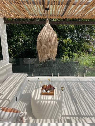 The image depicts a terrace in Buduk, Indonesia. A decorative lamp made of straw hangs from a wooden slat wall, which is constructed using dark wood. A glass railing separates the terrace from a lush green garden. Inside the terrace, the flooring is light-colored tile covered in shadows from an unseen structure. A low white sofa sits against the glass railing, and a round white table rests on the tile flooring. A wooden tray on the table holds cups and a red cloth. Next to the sofa is a cushion.