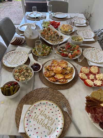 A long dining table, set for a celebratory meal, is presented from a top-down perspective in an indoor setting with a view of a patio through a large window. The table is covered with a marble-patterned tablecloth and adorned with various dishes, plates, and glassware, suggesting a birthday party or special occasion.
Several plates with a "Happy Birthday" design, featuring colorful confetti, are placed on woven placemats. To the left of the plates, stacks of sliced brown bread are visible, along with a tissue box. Various appetizers are spread across the table, including shrimp appetizers in small pastry shells, mini sandwiches with salmon and other toppings, and a salad composed of peas, carrots, potatoes, and ham. There is also a dish of sliced tomatoes topped with a creamy filling and a plate of cured meats and crackers. A small bowl of pickles and cherry tomatoes sits on a placemat.
Champagne flutes are placed at intervals around the table, some empty and some appearing to have contained liquid. A decorative spoon holds a dark red condiment, likely jam or sauce. The lighting suggests it is daytime, and the overall atmosphere is convivial and festive. The scene is set within a home in Dania Beach, Florida, United States of America, evidenced by the specific geographical location provided for this task. No people are visible in the image.