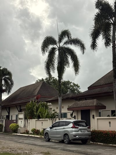 The image depicts a residential scene on a cloudy day in Ko Kaeo, Thailand. A silver Mitsubishi Xpander is parked on a paved road in front of a light-colored house with a dark brown tiled roof. The house is partially obscured by lush green foliage, including large-leafed plants and palm trees. A concrete wall with decorative elements runs along the property line, and there are various flowering plants in shades of red and green. The sky is heavily overcast with dramatic clouds. The road appears damp, suggesting recent rainfall. The overall atmosphere is one of a quiet, tropical suburban setting.