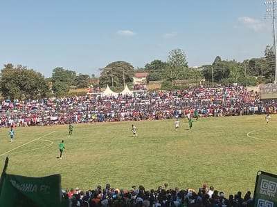 This wide shot captures an outdoor football (soccer) match in progress, viewed from a slightly elevated position within a stadium. The main activity is a game, with several players visible on the lush green grass pitch, distinguished by their green and white kits. A massive crowd, likely thousands, fills the tiered stands that encircle the field, packed tightly together and extending into the distance, suggesting high spectator engagement.
Beyond the densely populated stands, white tents, scattered buildings, and numerous trees are visible, framed by a bright, clear blue sky with only a few wispy clouds. A tall floodlight tower can also be seen on the right side above the stands. The sunny conditions clearly indicate daytime. In the immediate foreground, additional spectators are visible, along with parts of a green banner or flag on the left, displaying the partial text "...AHIA FC." On the right, a black and green sign with the logo and text "GO BANK" is prominently displayed. The scene depicts an energetic sporting event in Siaya, Kenya.