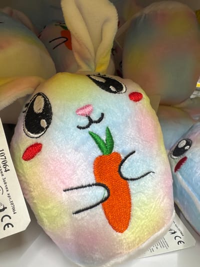 This is a close-up, slightly overhead shot of several colorful, plush bunny toys. The main bunny in focus is lying on its side, facing the viewer. It has a rainbow-colored fur pattern with pastel shades of pink, yellow, green, and blue. Its face is embroidered with large black eyes, a small pink nose, and a red stitched cheek. A prominent orange carrot with green leaves is embroidered on its face, and two black embroidered lines suggest a mouth or whiskers.

To the left and slightly behind the main bunny, another plush toy is visible. It appears to be a larger, rounder bunny with a similar pastel rainbow coloring, but its features are less clear. At the top right, a portion of another blue and white bunny toy is visible, also with embroidered facial features.

A white tag is partially visible in the bottom right corner, showing Cyrillic text and a "CE" mark. The text on the tag appears to be product information. The lighting is bright, suggesting an indoor setting like a store or a display. The overall impression is of a collection of soft, child-friendly toys.