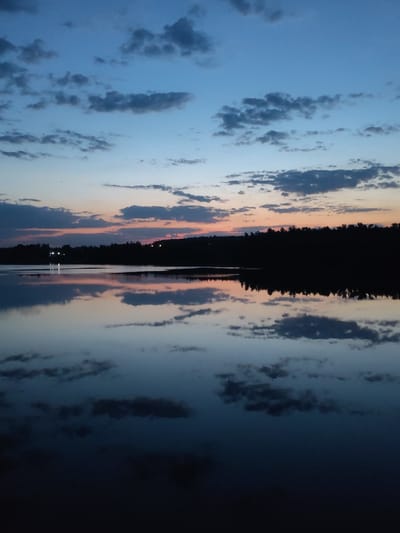 The image captures a twilight scene at a body of water, possibly a lake or a wide river, reflecting the sky. The sky displays a transition from blue at the top to shades of orange and pink near the horizon. Numerous clouds are scattered across the sky, mirrored in the water's surface. A dark treeline is visible along the horizon, with faint lights suggesting a distant settlement. The location is possibly Ermolino, Russia. The image depicts a tranquil, natural scene without the presence of people.
