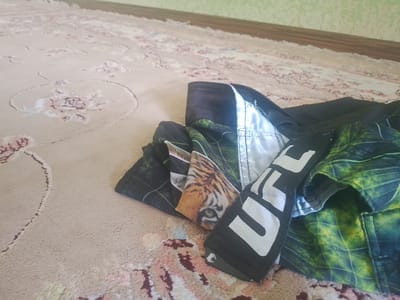The image shows a close-up of a pair of UFC shorts lying on a patterned carpet. The shorts are black with white lettering that reads "UFC" and have a green and black graphic print. A portion of a tiger's face is visible on one of the panels of the shorts. The carpet has a light beige background with intricate floral patterns in shades of pink, maroon, and cream. The background of the image shows a wall with light green textured wallpaper and a wooden baseboard. The scene is indoors, likely in a residential setting. There is no indication of the specific location within the city of Vahdat, Tajikistan.