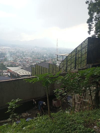 The image depicts a hillside overlooking the city of Gadog, Indonesia, under a hazy, overcast sky. In the foreground, dense green vegetation covers a steep slope. A concrete retaining wall runs horizontally across the mid-ground, below a large, dark, diagonally sloped mesh structure. At the base of the concrete wall, a single individual wearing a blue shirt and light-colored cap is engaged in manual labor, bending over and using a shovel in dark, wet soil. Nearby, a large, dark cylindrical container and several wooden planks or ladders are visible. A young papaya tree stands upright to the right of the worker. In the background, a sprawling urban area with numerous buildings and rooftops extends into the distance, with a tall communication tower prominent on the horizon and mountains obscured by haze further beyond.