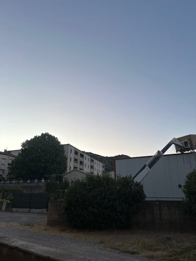 This image depicts a view of the city of Anduze, France, during what appears to be dusk or dawn, given the gradient of light in the sky. In the foreground, there is a gravel pathway leading towards a dark metal gate, flanked by lush green bushes on the right. Beyond the gate, a stone wall with a railing is visible, separating the foreground from a larger building complex.

Dominating the mid-ground are several multi-story buildings, characteristic of urban architecture. A particularly large, leafy tree stands prominently in front of one of these buildings. To the right, a corrugated metal structure is visible, topped with what appears to be a crane or lifting mechanism, with a light-colored box or container being held by its arm. Hills are visible in the background, suggesting the city's location within a more mountainous or hilly region. The overall atmosphere is tranquil, with the muted light and stillness of the scene.