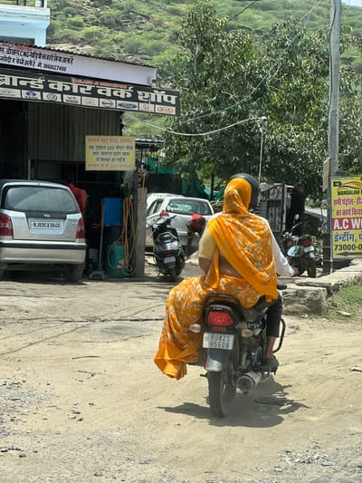 The image shows a dusty street scene in Ajmer, India, likely on a sunny day. In the foreground, a person wearing a bright orange and yellow floral sari with a matching headscarf is riding a black motorcycle. The person is facing away from the camera and appears to be the passenger, with their legs straddling the motorcycle. The motorcycle has a white license plate that reads "RJ42S H5608".
To the left of the motorcycle, a silver Hyundai Santro car is parked. The car has a license plate that reads "RJ09CA3840". Behind the car, an open-fronted workshop is visible with a sign that says "BRAND CAR WORK SHOP". The workshop appears to be under a slightly sloped roof. Inside the workshop, a person in a red shirt is partially visible.
Further down the street, a white car is parked next to a black motorcycle with a license plate that reads "RJ011 B5910". A street sign is visible to the right of the motorcycle, with Hindi text and the words "MANGALAM MOTORS".
The environment is characterized by unpaved, dusty ground with some scattered rocks and debris. In the background, a lush green hillside is visible, with trees and vegetation. Overhead, electrical wires are strung across poles, and a power line can be seen. The overall impression is of a busy, working-class area in a city with a warm climate.
