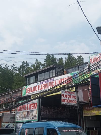 The image shows a shop named "Gemilang Plafon PVC & Interior," a distributor of PVC and interior materials. The storefront has a visible sign with contact numbers. The shop appears well-stocked with its products. Utility wires crisscross the foreground. A blue van is partially visible in front. Further background includes a line of tall green trees and a clear sky.