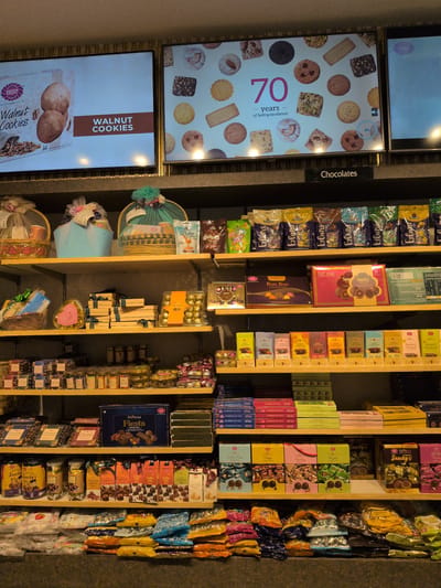 This image depicts a brightly lit interior of a confectionery or bakery store, likely located in Hyderabad, India, given the prominent "Karachi Bakery" branding. The scene is dominated by multiple wooden shelves showcasing a wide variety of packaged sweet products, with digital display screens above the main shelving unit. At the top, three horizontal digital screens are mounted. The left screen displays a "Karachi Bakery" advertisement for "Walnut Cookies," featuring an image of whole walnuts and cookies, with text indicating "Quality & Cookies since 1953." The central screen celebrates "70 years of baking excellence!" surrounded by a diverse array of cookies. The rightmost screen is partially visible, showing another cookie-related image. Below these screens, a black sign with white text reads "Chocolates." The store's main display consists of six wooden shelves, densely packed with items. The topmost shelf on the left features three elaborately wrapped gift baskets: one in gold, one in light blue, and one in teal, each adorned with ribbons and decorative elements. To the right of these baskets, several pouches of snacks and sweets are visible, including "Butter Bites" and "Coffee Toffees." The right half of this shelf is lined with multiple colorful bags of "Sapphire Eclairs" in purple, blue, and green packaging. The second shelf from the top on the left side holds a brown woven basket wrapped with green ribbon and a heart-shaped golden box with a pink ribbon. The middle section of this shelf contains stacks of rectangular gift boxes, possibly chocolates, tied with dark ribbons. On the right, a clear box labeled "Snacky's" contains spherical chocolates, reminiscent of Ferrero Rocher. Also present are a purple box labeled "Bon Bon Collection" and a pink box with "Coco Atini" and "Snacky's" branding. The third shelf features a long, organized row of small "Coco Atini" chocolate bars, each displaying a different cocoa percentage (e.g., 50% Dark, 55% Dark, 66% Dark, 78% Dark, 49% Milk, 38% Milk) and various flavors like "Roasted Almond" and "Roasted Hazelnut," packaged in colorful wrappers. The fourth shelf showcases numerous clear jars filled with assorted chocolates and candies, many bearing the "Coco Atini" label. A prominent blue box in the center is labeled "Coco Atini Fiesta Collection." Additional rectangular boxes and chocolate products are arranged on either side. The fifth shelf from the top displays various "Snacky's" branded products, including jars and packets, some marked with a "₹ 10" price point. Adjacent