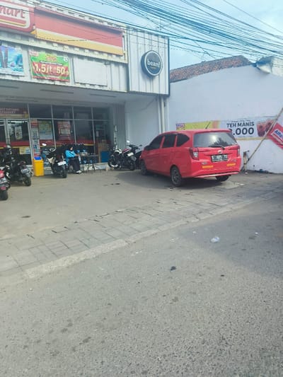 The scene is in front of a store in Purwakarta, Indonesia, with coordinates -6.546 latitude and 107.442 longitude. A red car is parked facing the street. Two motorcycles are parked on the sidewalk. A person wearing a blue hijab and a white shirt is seated on the sidewalk with a yellow bag in front of them. The store front is white with red signage. "Bean!" is written on the front of the building. The road is paved, and the sidewalk is paved with gray bricks. The sidewalk is separated from the road by a curb. There are many power lines in the sky above the scene. The street is in a residential area. There are signs on the building behind the car, advertising "Teh Manis 3,000" and "Alfamart." There is a yellow bin on the sidewalk. The street is empty.