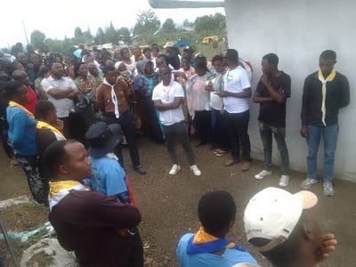 A diverse crowd gathers outdoors, mostly adults, near a building. Many wear casual clothing, while some individuals stand out with scout-like uniforms and neckerchiefs in various colors. A man in a white t-shirt with crossed arms appears to be addressing the group. The ground is dirt and grass, with trees and a cloudy sky in the background. The atmosphere suggests a community event or meeting.