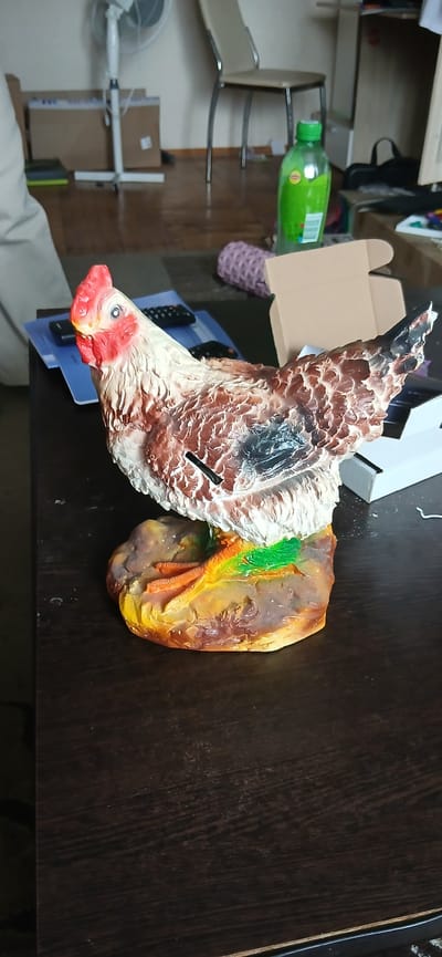 The image displays a decorative ceramic chicken figurine positioned on a dark wooden table. The figurine is detailed, with visible feathers painted in shades of brown, white, and black, and a red comb and wattles. It stands on a base that resembles rocks and grass, with orange bird feet showing. In the background, a white pedestal fan is visible, along with cardboard boxes, a chair, and a green plastic bottle. The scene appears to be indoors, possibly a room in a residential setting. There is no information in the image to indicate the location is Yaransk, Russia.