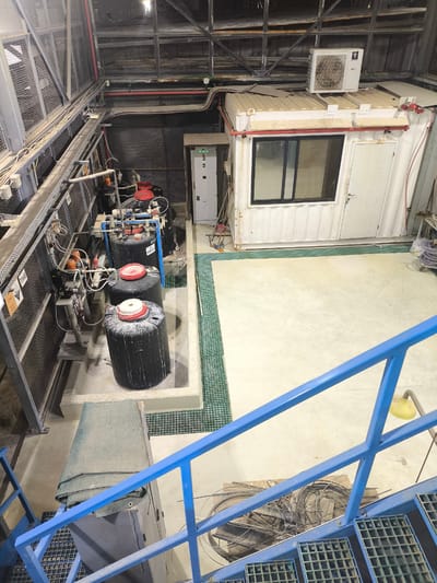 The image shows an interior view of an industrial facility in Dimona, Israel. The scene depicts a processing area with several large, black cylindrical tanks, some with red lids, arranged in a concrete containment area. Pipes and electrical conduits are visible, connecting the tanks to machinery and a control panel. A white, modular building with a window and door, likely an office or control room, is situated to the right. An air conditioning unit is mounted on its roof. Various industrial equipment, including pumps and wiring, are present. The foreground shows a blue metal staircase with a mat on a metal platform. The overall environment appears to be a manufacturing or processing plant, with elements of dust and wear evident on surfaces, suggesting ongoing industrial operations.