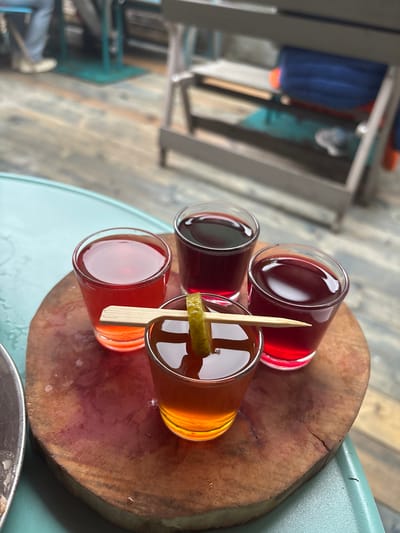 The image shows a wooden cutting board with four small shot glasses of various colored liquids. A small piece of green garnish, possibly a lime or cucumber slice, is speared by a wooden pick and placed across the front shot glass. The shot glasses are arranged in a circular pattern on the board. The background appears to be an outdoor setting with wooden planks on the ground and blurred furniture. No specific location in Komarovo, Russia is identifiable.
