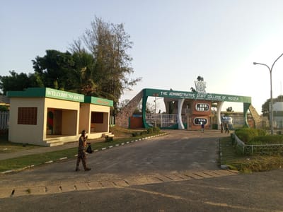 The image shows the entrance to the Administrative Staff College of Nigeria (ASCON) in Tawpo, Badagry, Lagos State, Nigeria. A man dressed in colorful patterned attire walks across the road in front of the entrance gate, carrying a black bag. Behind him to the left is a building with a sign that says "WELCOME TO ASCON Since 1972". The entrance gate is green and white, with "THE ADMINISTRATIVE STAFF COLLEGE OF NIGERIA P.M.B. 1004 TOPO BADACRY LAGOS STATE NIGERIA" written on it. Several other people are seen standing near the gate. There are trees and bushes around the entrance. A street light is visible on the right side of the road. The sky is bright with morning light.