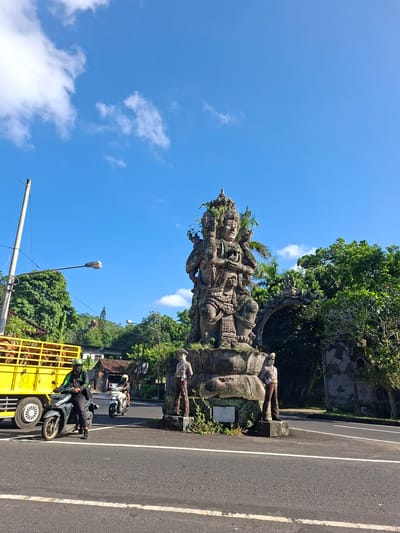 In Banjarangkan, Indonesia, a street scene unfolds under a bright blue sky dotted with white clouds. Dominating the center of the frame is a large, weathered stone statue, likely depicting a deity, adorned with multiple arms and intricate headdresses. The statue sits on a tiered stone base, with patches of moss and vines growing on it. Two smaller stone figures, one resembling a police officer in uniform and the other appearing to be a woman, stand at the base of the statue, one on each side.
To the left, a bright yellow truck filled with cattle is stopped at the roadside. A person wearing a green helmet and dark clothing rides a motorcycle in front of the truck, with another person on a white motorcycle behind them. A lamppost stands tall to the left of the truck, and another lamppost is visible further down the road. Lush green trees line the background, with hints of buildings and structures behind them. The road in the foreground is paved and marked with white lines. The overall atmosphere is that of a bustling street in a tropical location.
