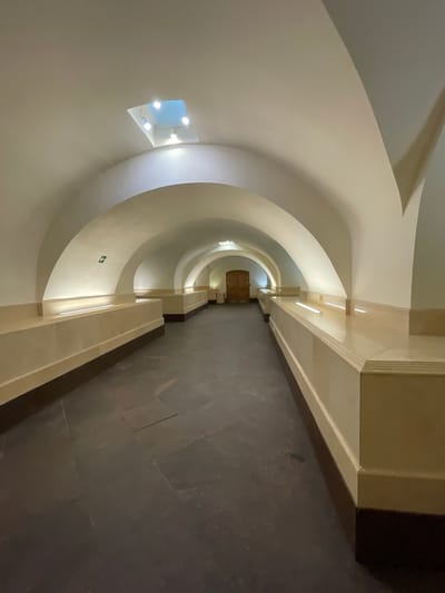 The image shows a long, arched corridor with a dark tiled floor. The walls are a light, creamy color, and there are decorative niches with lighting along the sides. A skylight with several recessed lights is visible in the ceiling of the first arch. The corridor extends into the distance, where a wooden door is visible at the far end. An illuminated green arrow sign indicating direction is mounted on the wall to the left. This scene is located in Strelna, Russia, specifically within the Konstantinovsky Palace complex.