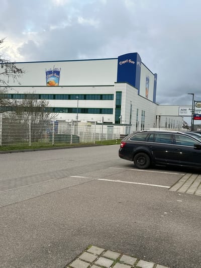 The image displays an exterior view of the Capri-Sun production facility located on Rudolf-Wild-Straße in Eppelheim, Germany, under an overcast sky. The primary subject is a multi-story industrial building featuring a white and blue facade. A prominent blue section on the building's corner displays the name "Capri-Sun" in white lettering. A large graphic of a "Capri-Sun Orange" pouch is affixed to a white section of the building's upper wall. Lower levels of the building exhibit multiple horizontal rows of windows with green frames. A covered walkway or bridge extends from the right side of the main structure.

In the foreground, an asphalt parking area is visible, delineated by white painted lines. A dark-colored station wagon, fitted with black steel wheels, is parked on the right side of the frame, with its rear section and passenger side visible. A band of grey paver stones traverses the asphalt surface in the mid-ground. A low, grey chain-link fence separates the paved area from a narrow strip of ground featuring sparse, leafless deciduous trees and bushes. In the background, a pole-mounted road sign assembly is visible, displaying text that includes "ADM Anlieferung" and "Capri-Sun." No individuals are present in the scene. The sky is uniformly cloudy.