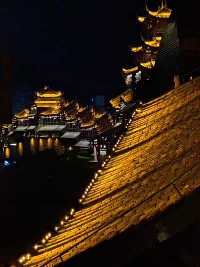 The image shows illuminated buildings at night in Zhuoshui, China. A large bamboo structure with strung lights is positioned at the top right. Multiple buildings are visible along a dark body of water on the left side of the image. The buildings have ornate rooflines. Reflections of the lights are visible in the water.