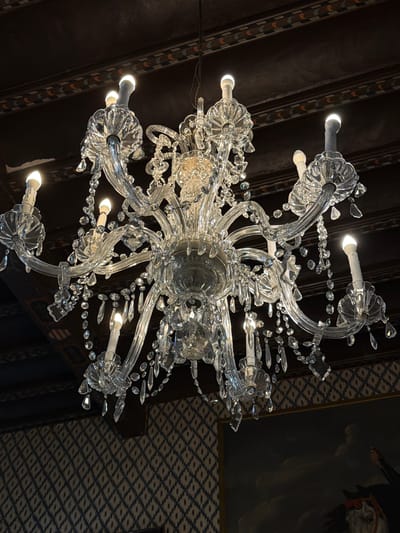 An angled close-up depicts a clear glass chandelier hanging from a dark-toned ceiling. The chandelier features multiple curved arms adorned with small glass crystals, and small light bulbs are visible at the ends of some arms. The ceiling is dark, possibly wood, with a decorative pattern along its beams. In the background, a corner of a painting with a dark subject is visible against a wall with a blue and white geometric pattern. The scene does not show any people. There are no indications that the image was taken in Bunyola, Spain.