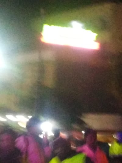 A blurry, low-quality image captures a crowd at night. Several people are visible in the foreground, their features obscured by motion blur and poor lighting. A glowing, rectangular sign with green and pink hues is seen in the upper part of the image. The dark background further contributes to the overall indistinctness, making details hard to discern.