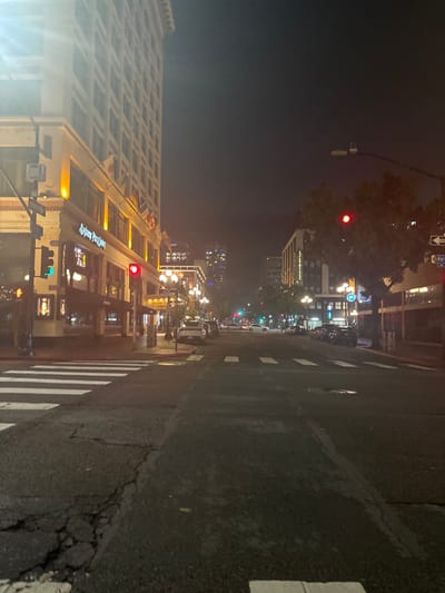 A nighttime street view reveals a city with lit buildings, streetlights, and traffic signals. A large building with a grid-like facade flanks the left side. The street's surface shows cracks and wear, with crosswalk markings visible. Further down, more buildings and lights add to the urban ambiance. The sky is dark, and a slight haze softens the distant lights.