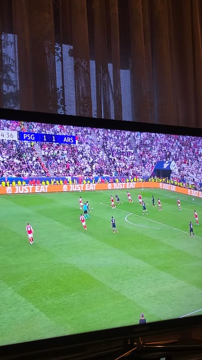 The image shows a television screen displaying a soccer match. The score is PSG 1, ARS 1. The image is rotated counter-clockwise. The match appears to be in progress, with players visible on a green field. Spectators fill the stadium in the background. "JUST EAT" is displayed on the side of the field. The TV is located indoors, in front of window blinds. There is no evidence of a location in Rogachevo, Bulgaria.
