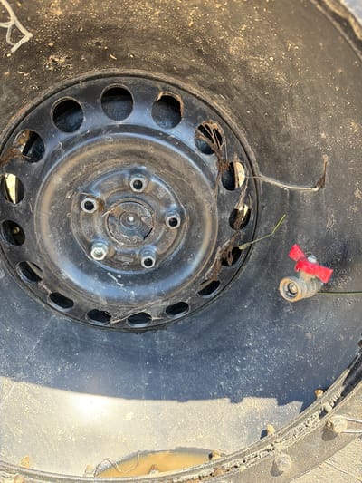 This is a close-up, forensic image of a damaged tire rim with a brass valve stem fitting attached to it. The rim is black and appears to be made of steel. It is covered in dirt and debris, including what looks like dried grass or straw. There are several lug nut holes visible around the center of the rim. A small brass valve stem fitting with a red lever handle is attached to the rim on the right side of the image. The fitting appears to be a ball valve. The tire itself is partially visible at the bottom of the frame and appears to be flat and damaged. The background is blurred and out of focus. The image does not contain any contextual information that would indicate its location in the city of Аур, Russia, or any other specific location. There are no people, actions, or interactions depicted in the image. The scene is one of abandonment or disuse, with the damaged tire and valve stem suggesting a discarded or neglected object.