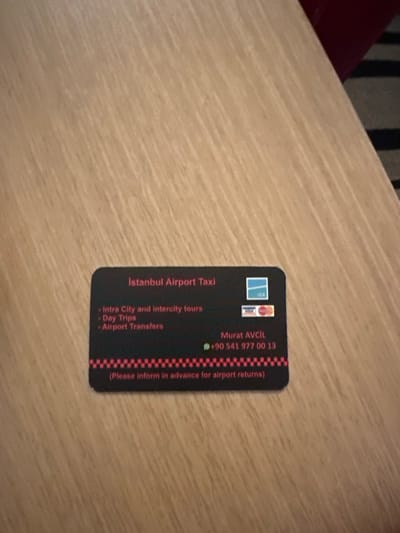 The image displays a black business card with rounded corners, centrally placed on a light-toned wooden surface featuring a distinct diagonal grain. The card advertises "Istanbul Airport Taxi" in red text at the top. Listed services in red include "Intra City and Intercity tours," "Day Trips," and "Airport Transfers." Payment method logos for IGA, Visa, Mastercard, and an additional card are visible on the right side. The contact information "Murat AVCil" and the WhatsApp number "+90 541 977 00 13" are provided. A red and black checkered pattern precedes the instruction "(Please inform in advance for airport returns)" at the bottom. The wooden surface, possibly a table, fills the majority of the frame. A blurred dark red object and a striped grey and black textile, likely a carpet or upholstery, are visible in the upper right background, indicating an indoor setting. No individuals, actions, or direct interactions are depicted. The scene is located in Zeytinburnu, Turkey.