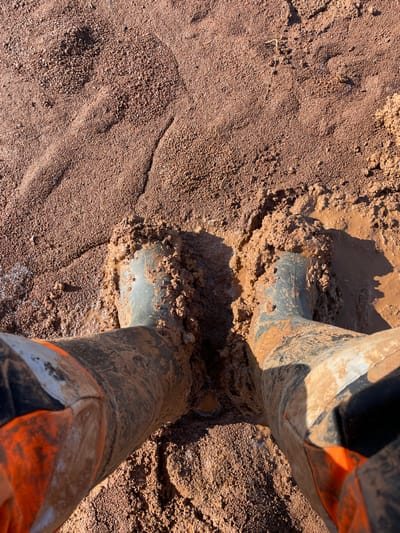 A first-person, downward-angle perspective captures two feet encased in dark green, mud-covered rubber boots, submerged in reddish-brown, viscous mud. The boots are heavily encrusted with wet mud extending above the ankles. Portions of orange, potentially water-resistant, trousers are visible immediately above the boot shafts. The surrounding ground is composed of reddish-brown fine-grained soil and granular material, interspersed with areas of standing muddy water and deeper mud, particularly around the boots. Distinct shadows are cast to the right, indicating direct sunlight from the upper-left. Small, irregular patches of white material, possibly crystalline deposits, are present on the mud surface towards the upper-left. The scene depicts a person stationary or having recently moved through challenging muddy terrain. The specific location within Gari, Russia, is not visually identifiable.