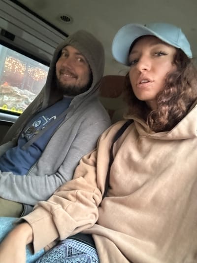 Two people are in a vehicle. The person in the front is wearing a light blue baseball cap and a tan hoodie. The person in the back is wearing a gray hooded sweatshirt and a blue t-shirt. It is daytime and appears to be raining outside. The exact location in Binh Lu, Vietnam cannot be determined from the image.