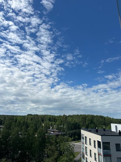The image shows a view from a high vantage point, looking out over a treeline towards a bright blue sky filled with scattered white clouds. In the foreground, a portion of a modern apartment building is visible, with white brickwork and dark window frames. The building extends out of the frame to the right. Below the apartment building and the treeline, there appears to be a suburban landscape with houses and roads. The overall scene suggests a clear day in a developed area surrounded by nature.