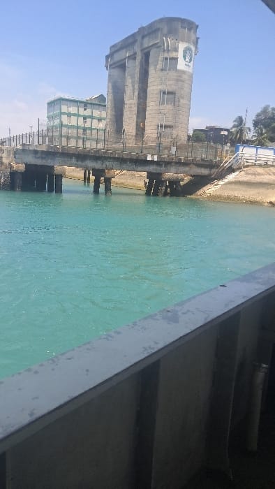 The image depicts a daytime scene at the Likoni Channel ferry crossing in the Ganjoni/Shimanzi ward, Kenya. The foreground features the upper grey railing of a vessel, from which the photograph was taken. Beyond the railing, calm, turquoise-colored water fills the lower-mid frame.
Dominating the midground and right-mid frame is a large, weathered concrete structure, identifiable as part of the Likoni Ferry terminal infrastructure. This structure is tower-like, with a rounded top and prominent vertical recesses, appearing to be a control or operational building. A green and white circular logo is affixed to its side. Adjacent to and extending from this structure is a concrete pier or bridge supported by multiple concrete pillars submerged in the water. The pier features a dark metal railing along its edge, with some chains visible hanging into the water. On the right, beneath the pier, a rocky and sandy shoreline is visible.
In the background to the left, a multi-story building, light green with numerous windows, stands. The sky above is clear blue, indicating bright daylight conditions. No individuals are visible, and no specific actions or interactions are depicted within the scene.