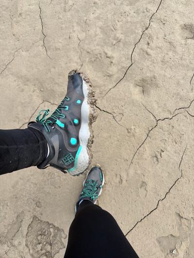 The image shows two feet, one clad in a muddy black and gray Columbia hiking boot with bright teal laces, and the other in a similar gray sneaker with teal laces, both partially covered by black leggings. The shoes are positioned on a cracked, dry, light brown, earthy surface that appears to be mudflats or a dried-up riverbed. The cracked texture of the ground is prominent, with intricate lines creating a mosaic-like pattern. The context provided mentions the city of Sauris / Zahre, Italy, but this is not visually evident from the image itself. The lighting suggests an outdoor, possibly overcast day.