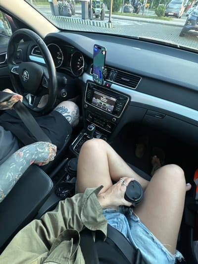 The image shows the interior of a car, likely a Skoda, with two people inside. The driver, a man with extensive tattoos on his arms, is wearing black shorts and a grey t-shirt, and is seen fastening his seatbelt. The passenger, a woman with ripped denim shorts, a green jacket, and multiple rings on her fingers, is holding a coffee cup. The car's dashboard shows a navigation app on a phone mounted on the windshield. Outside the car, a paved lot is visible with other vehicles, including a grey sedan and a white SUV, suggesting the car is parked in a public area. The background suggests an urban or suburban setting, possibly a parking lot or street in Barvikha, Russia, indicated by the presence of trees, buildings, and streetlights. The lighting suggests it is daytime, possibly late afternoon due to the warm glow.