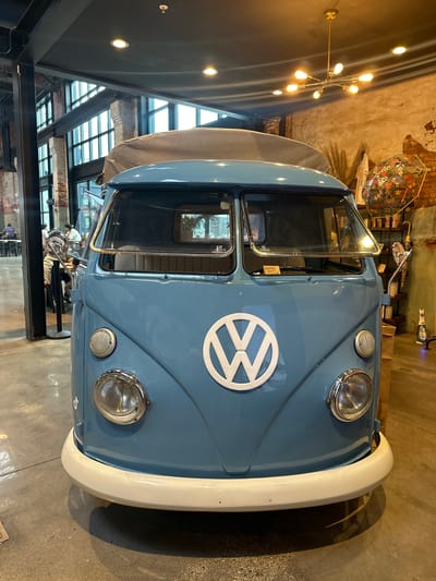 The image shows the front of a light blue Volkswagen van, likely a vintage model, parked indoors. The van features a white VW logo on the hood and a white front bumper. The setting appears to be a restaurant or retail space with large windows and a modern architectural style. A distinctive chandelier is visible in the background, along with other decorative items. Other people can be seen walking near and behind the van. The photo was taken at Heights Public Market at Armature Works.