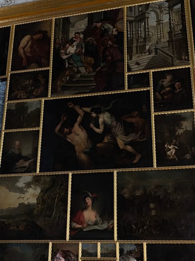 The image shows a collection of oil paintings displayed in ornate gilded frames, arranged in a grid-like pattern. The paintings depict various scenes, including religious subjects, mythological figures, genre scenes, and landscapes. Some of the notable paintings include:

*   A painting on the top left depicts a muscular male figure reclining and holding a chalice.
*   To its right, a group of figures is shown in a dramatic scene, possibly religious, with women in flowing robes and men in darker attire.
*   The top right painting is an architectural scene featuring a grand staircase and classical structures, with small figures scattered throughout.
*   Below these, a large central painting dominates the composition. It shows two male figures in a struggle; one is bound and appears to be in pain, while the other is holding a whip or rope.
*   To the left of the central painting, there are two smaller rectangular paintings. The upper one shows a dark landscape with a cluster of fruits or flowers. The lower one depicts an elderly man with a white beard seated at a desk, reading a book.
*   To the right of the central painting, there are two more rectangular paintings. The upper one shows two individuals, one pouring liquid from a jug into a bowl held by the other. The lower one depicts a scene with a nude female figure in a wooded landscape, with other figures in the background.
*   Below the central painting, there is a horizontal painting of a landscape with rolling hills, a body of water, and trees.
*   To the right of this landscape, there is a portrait of a woman wearing a red garment and a feathered hat, looking down at a book.
*   Further to the right, a large painting depicts a dark, crowded scene with architectural elements and numerous figures, possibly a battle or a chaotic event.

The paintings are arranged within a larger gilded frame, suggesting they are part of a decorative wall panel. The overall impression is of a rich and diverse collection of artworks, likely housed in a museum or a grand residence. The specific location in Alexandrovskaya, Russia, is not visually evident from the paintings themselves.