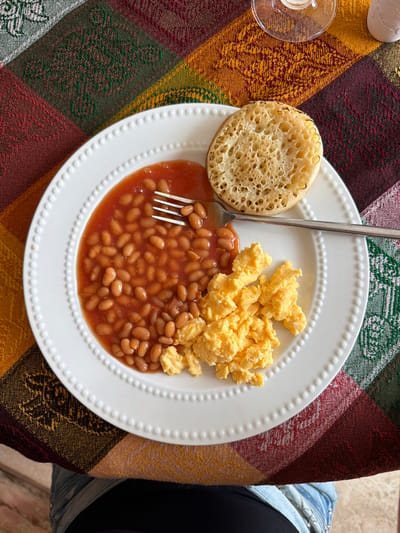 A high-angle, close-up shot captures a white, round plate filled with a breakfast meal. The plate is centered on a colorful, woven tablecloth with a pattern of horizontal stripes in shades of burgundy, yellow, green, and brown.
The meal on the plate consists of a generous portion of baked beans in a tomato sauce, a pile of scrambled eggs, and a single, round crumpet. A silver fork rests on the plate, partially submerged in the beans. To the left of the plate, a clear glass dish with a small amount of liquid is visible.
In the background, the edge of a person's dark clothing and a denim jacket are visible, suggesting the plate is on a table. The lighting indicates it is daytime, and the overall scene conveys a casual breakfast setting. No visible text is present in the image.