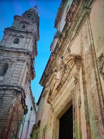 A low-angle shot captures a tall, weathered stone bell tower on the left, against a clear blue sky. The tower features multiple levels with arched windows and intricate architectural details, showing signs of age and possibly some overgrowth. To the right of the tower, the facade of another building is visible, also made of stone with ornate carvings and columns around an entrance. This building appears to be lighter in color and also shows considerable age. The perspective emphasizes the grandeur and historical nature of these structures.
Based on available information, this scene is likely the St. John of Nepomuk Cathedral (also known as the Roman Catholic Cathedral of St. John of Nepomuk), located in Zrenjanin, Serbia.